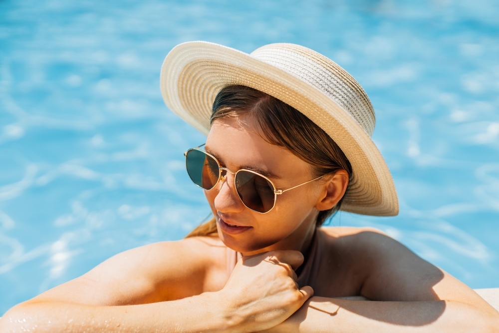 Beautiful woman in a hat and sunglasses relaxing in the water of the pool. A girl with healthy tanned skin, a gorgeous face, and wet hair enjoys the summer sun on a hot summer day at the edge