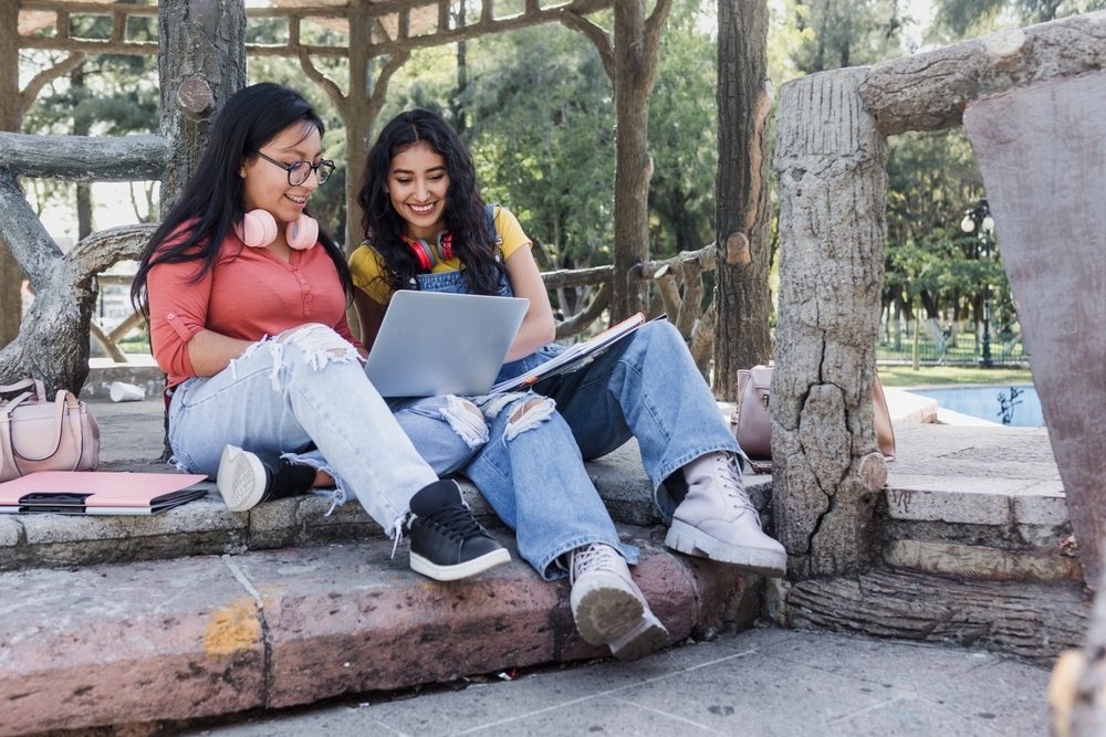 two young latin women students using laptop sitting on bench park in Mexico Latin America, hispanic girls studying 