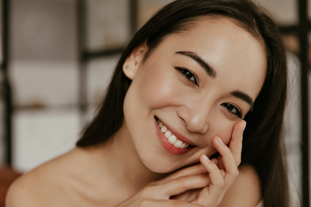 Happy young woman smiles sincerely. Attractive brunette Asian lady touches gently her face skin and looks into camera in good mood.