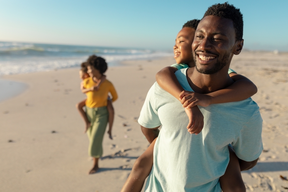 Happy african american man piggybacking son with mother and daughter walking in background at beach. unaltered, family, lifestyle, togetherness, enjoyment and holiday concept.