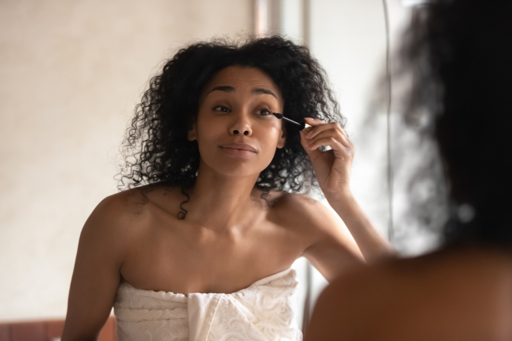 Beautiful young african American woman in towel look in mirror in bathroom paint eyelashes with black thickening mascara, biracial female get ready do makeup in home bath, beauty procedure concept