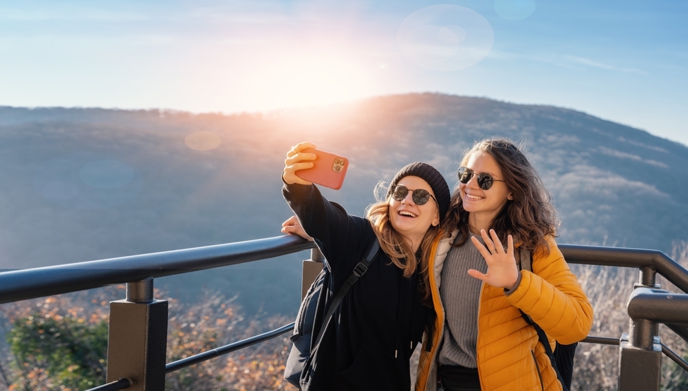 Two happy cheerful young girls in sunglasses traveling together taking selfie on smartphone