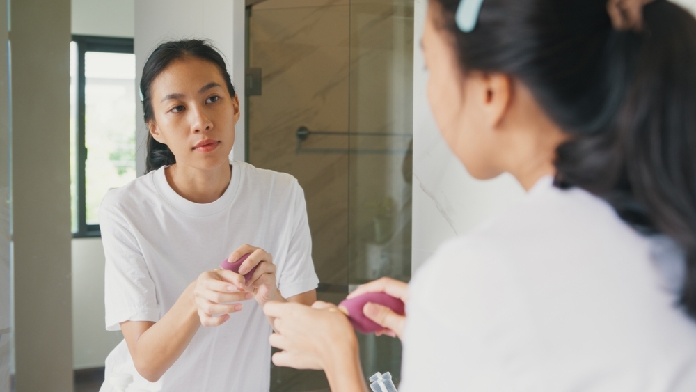Young Asian happy woman looking at her reflection in mirror use makeup sponge apply foundation on her face in bathroom. Lifestyle and people, Natural Skin Care Cosmetics, Morning routine concept.