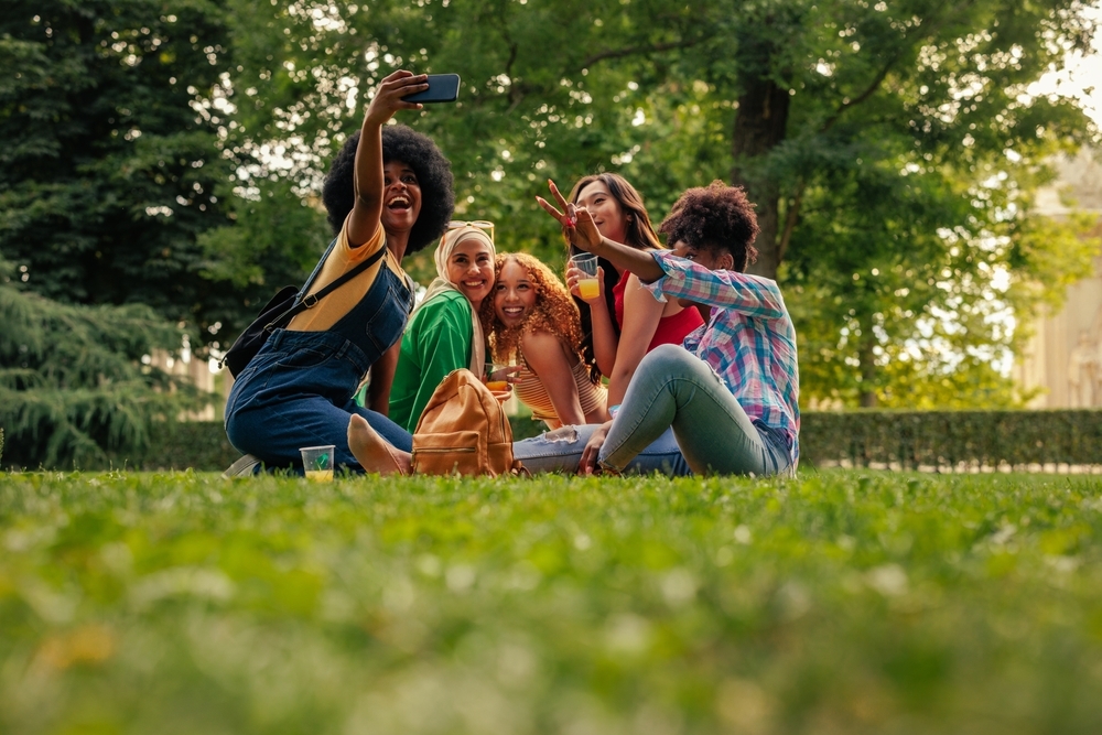 A small cheerful party of multiethnic girls are on the lawn in the city park taking a selfie with a cellphone.