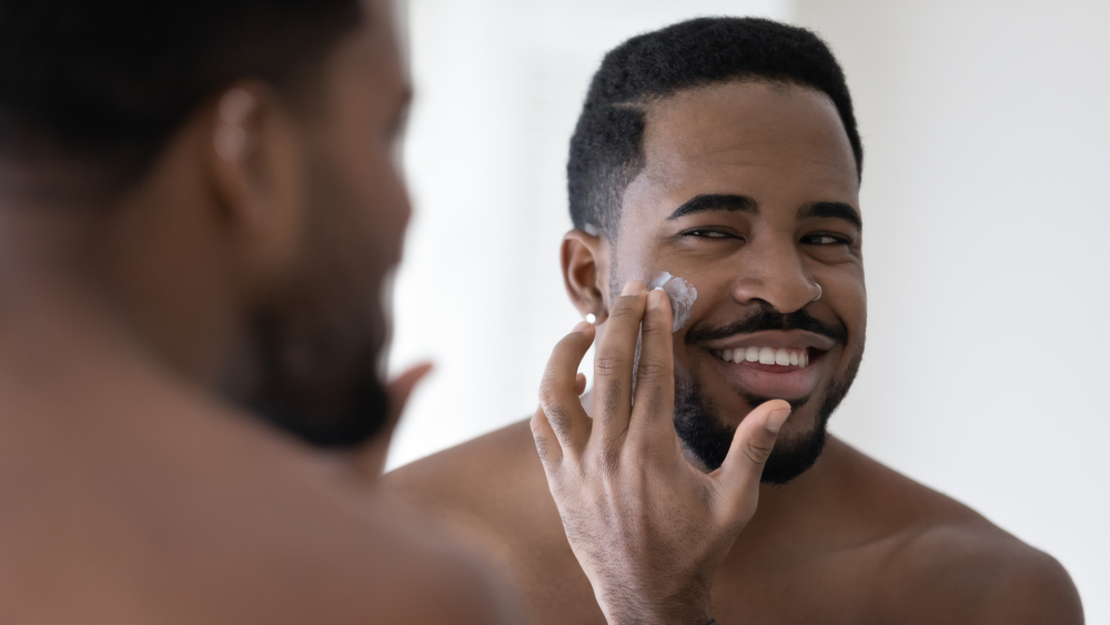 Happy handsome African American man applying moisturizer on face, putting collagen anti age cream, sunscreen, cleansing lotion, caring for hydrating soft skin, reducing wrinkles, acne, eyebags