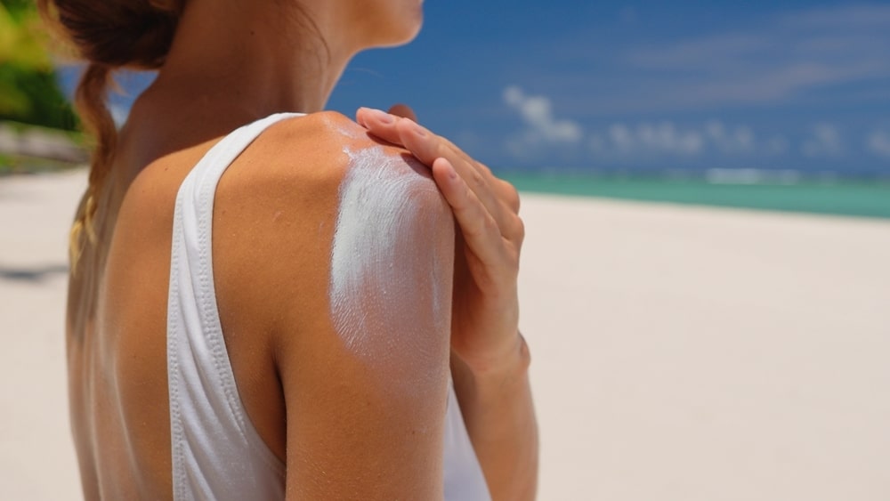 Close up of a happy smiling young woman is applying a sunscreen or sun tanning lotion on a shoulder to take care of her skin on a seaside beach during holidays vacation.