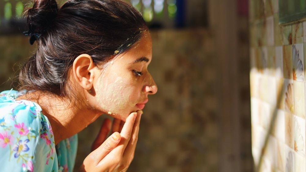 A beautiful woman is washing facial mask in bathroom after applying face mask. Girl spraying water on her face standing in front of mirror at home. mud mask or homemade multani mitti mask.