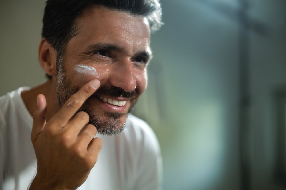 An young smiling man is applying after cleaning his face day or night cream to take care of skin behind a mirror in a bathroom.